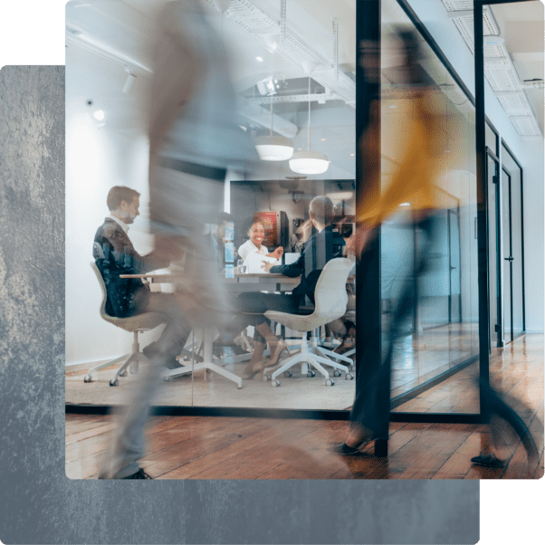 Blurred figures walking past a glass-walled conference room where a team is having a meeting.
