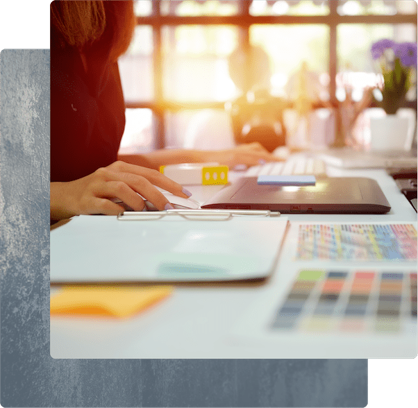 A designer working at a desk with a computer mouse, tablet, and color swatches in a sunlit office.
