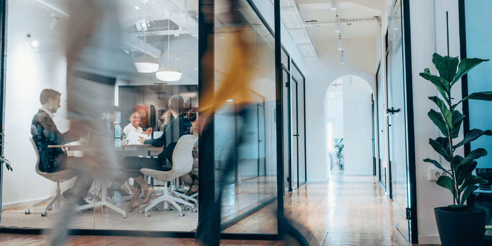 Business professionals in motion passing a glass-walled conference room where a team meeting is in progress in a modern office hallway.