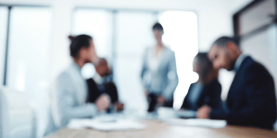 Blurred background of business professionals gathered around a conference table during a presentation or meeting.
