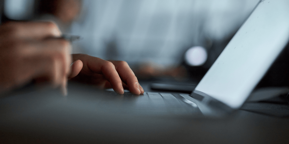 Close-up of a hand using a laptop trackpad with a pen in the other hand in a dimly lit workspace.