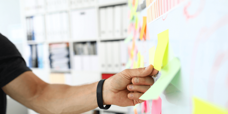 A person placing colorful sticky notes on a whiteboard covered in project planning notes in an office environment.