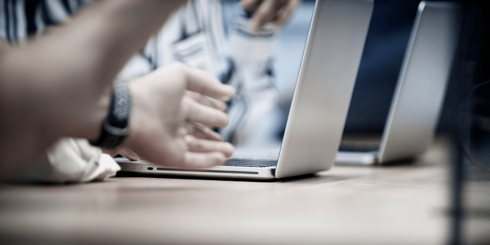 Close-up of hands typing on a silver laptop at a table with additional laptops visible in the background.