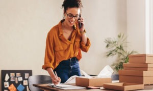 Woman on phone standing at a desk writing on papers.